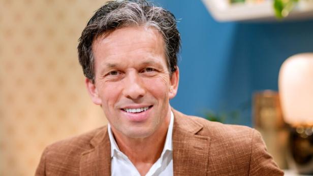 Ein Mann mit braunem Sakko und weißem Hemd sitzt lächelnd auf einem Sofa in einem modern eingerichteten Raum.