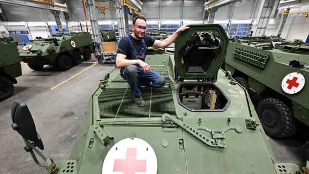 Ein Mann kniet auf einem grünen Panzer mit rotem Kreuz und geöffneter Luke in einer Werkhalle.