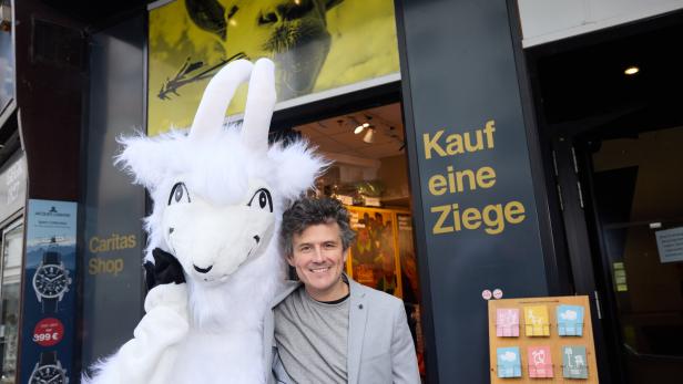 Ein Mann lächelt neben einem Ziegen-Maskottchen vor einem Laden mit der Aufschrift "Kauf eine Ziege".