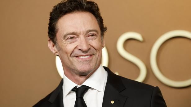 Ein Mann im schwarzen Anzug und schwarzer Krawatte lächelt vor einem braunen Hintergrund mit großen Buchstaben.