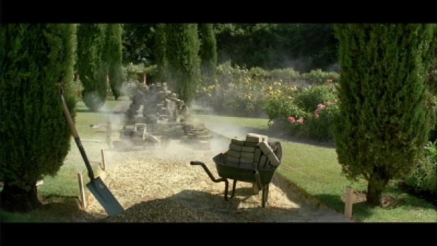 Eine Schubkarre voller Steine steht in einem Garten mit Zypressen und Blumen.