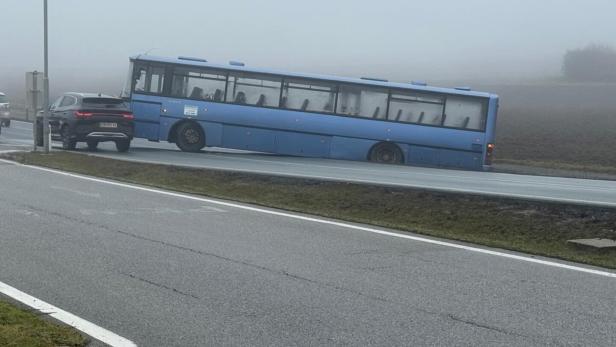 Ein blauer Bus steckt schräg im Straßengraben, während Autos auf einer nebligen Straße vorbeifahren.