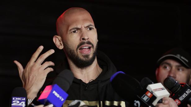 Ein Mann mit Bart spricht gestikulierend vor mehreren Mikrofonen, im Hintergrund steht eine weitere Person.