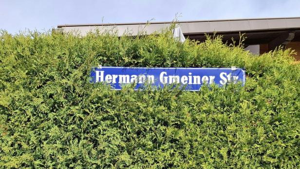 Ein blaues Straßenschild mit der Aufschrift „Hermann Gmeiner Str.“ ist in einer grünen Hecke befestigt.