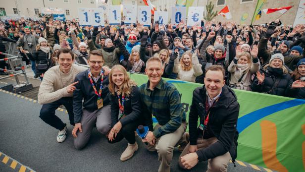 Fünf Personen knien vor einer jubelnden Menschenmenge, die Schilder mit der Spendensumme 151.340 € hochhält.