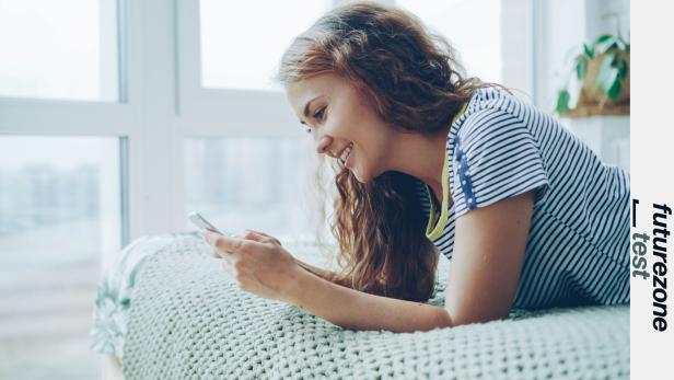 Junge Frau liegt lächelnd auf dem Bett und nutzt ihr Smartphone vor einem großen Fenster.