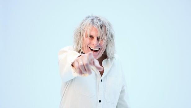 Eine Person mit langen, blonden Haaren lacht und zeigt mit dem Finger nach vorne, trägt helle Kleidung und hält eine Brille in der Hand.