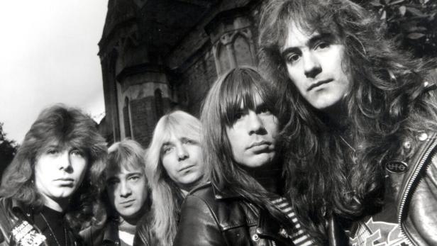 Fünf Männer mit langen Haaren und Lederjacken stehen vor einem alten, dunklen Gebäude.