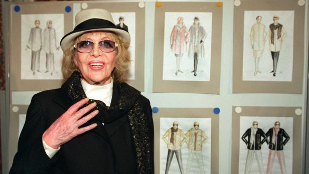 Eine ältere Frau mit Hut und Sonnenbrille steht lächelnd vor einer Wand mit mehreren Modezeichnungen.