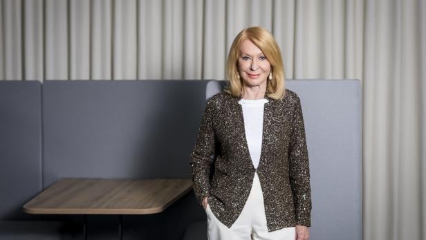 Eine Frau mit blondem Haar trägt eine glitzernde Jacke und weiße Hose, steht vor grauen Sitzmöbeln und hellem Vorhang.