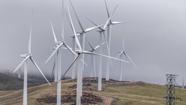 Mehrere Windräder stehen auf grünen Hügeln unter einem bewölkten Himmel, rechts ist ein Strommast zu sehen.