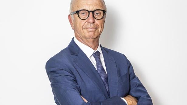 Älterer Mann mit Brille und Glatze trägt blauen Anzug und steht mit verschränkten Armen vor weißem Hintergrund.