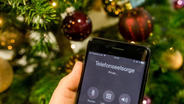 Eine Hand hält ein Smartphone mit einem Anruf bei der Telefonseelsorge vor einem geschmückten Weihnachtsbaum.