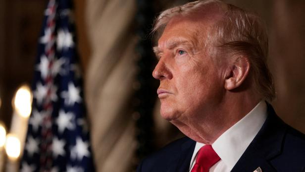 Ein Mann im Anzug mit roter Krawatte blickt ernst zur Seite, im Hintergrund ist eine US-Flagge zu sehen.