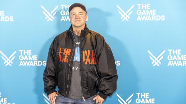Ein Mann mit schwarzer Jacke und Basecap steht vor einer blauen Wand mit „The Game Awards“-Logos.
