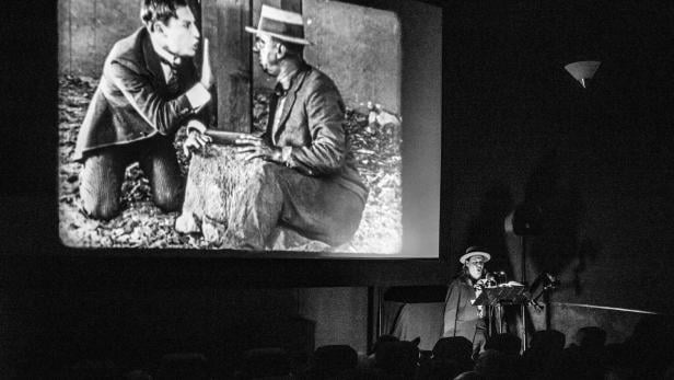 Eine Person spielt live Musik vor Publikum, während ein Stummfilm mit zwei Männern gezeigt wird.