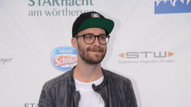 Ein Mann mit Brille, Kappe und Lederjacke steht vor einem Werbebanner.