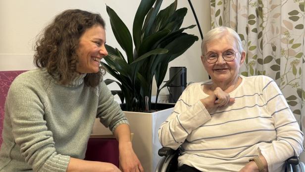 Eine Frau mit lockigen Haaren unterhält sich lächelnd mit einer älteren Dame im Rollstuhl.