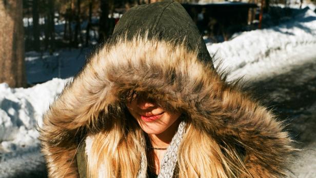 Eine Frau trägt eine Winterjacke mit Fellkapuze im Schnee, wobei das Fell außen sichtbar das Gesicht vor Kälte schützt.