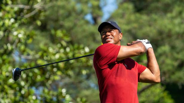 Ein Mann mit rotem Shirt und schwarzer Kappe schwingt einen Golfschläger auf einem grünen Golfplatz.