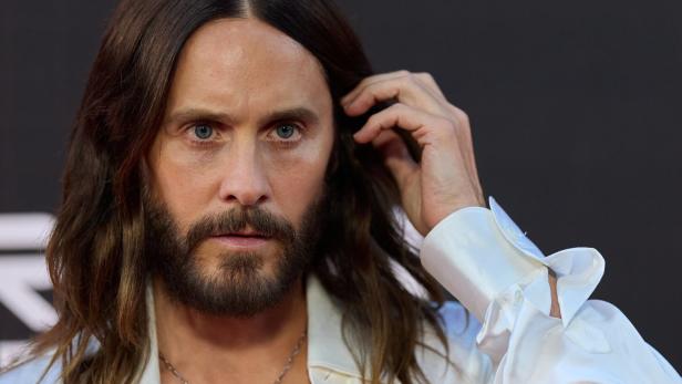 Jared Leto mit langem Haar und Bart trägt ein weißes Hemd und blickt direkt in die Kamera.