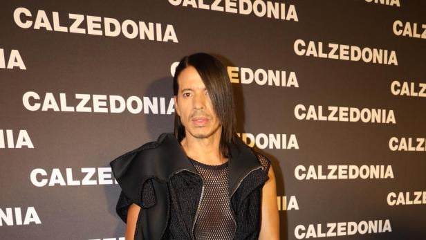 Eine Person mit langen dunklen Haaren trägt einen schwarzen, auffälligen Einteiler und Plateauschuhe vor einer Calzedonia-Wand.