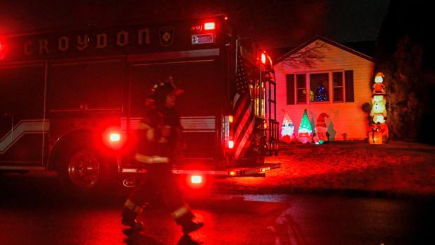 Ein Feuerwehrfahrzeug mit blinkenden Lichtern steht vor einem weihnachtlich geschmückten Haus, ein Feuerwehrmann läuft vorbei.