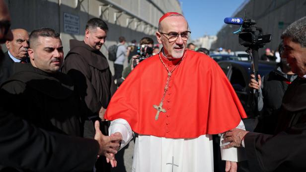Ein Kardinal in rotem Umhang wird von Menschen begrüßt, während Kameras die Szene festhalten.