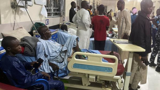 Mehrere Menschen befinden sich in einem vollen Krankenhauszimmer, einige liegen in Betten, andere stehen oder sitzen.