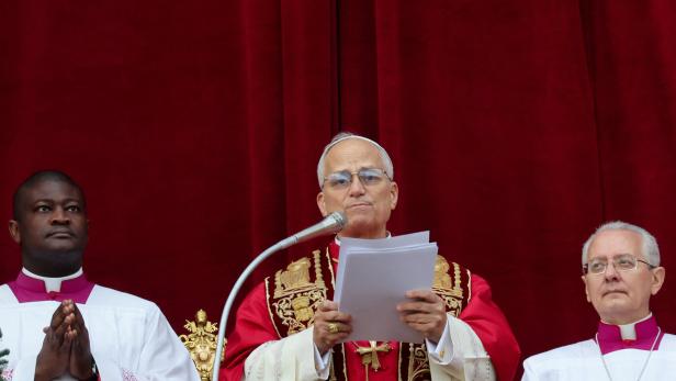 Ein Geistlicher in prunkvoller liturgischer Kleidung hält eine Rede am Mikrofon, flankiert von zwei weiteren Geistlichen.