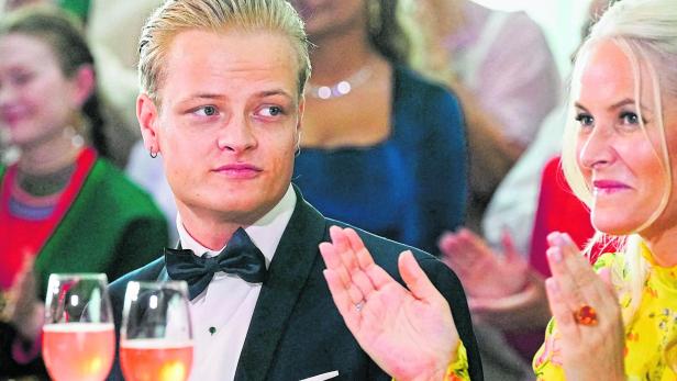 Ein junger Mann im Smoking sitzt neben einer Frau mit blondem Haar und gelbem Kleid, beide wirken festlich und aufmerksam.