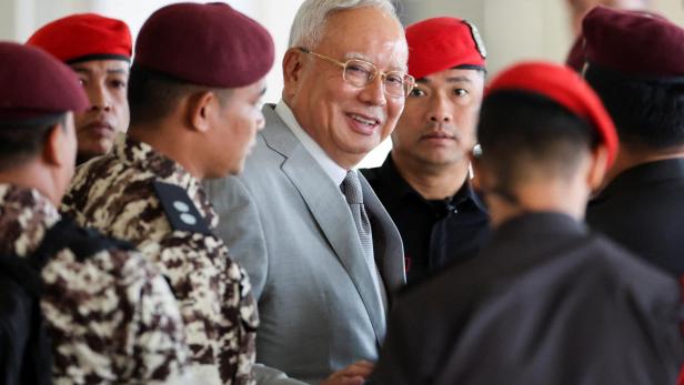 Ein älterer Mann im Anzug wird von mehreren uniformierten Männern mit roten Baretten begleitet.