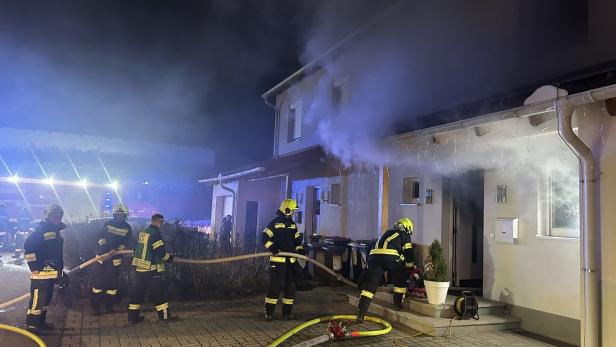 Mehrere Feuerwehrleute bekämpfen nachts einen Brand in einem Wohnhaus, aus dem dichter Rauch dringt.
