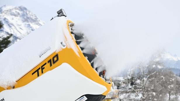 Eine gelbe Schneekanone bläst Schnee in eine winterliche Landschaft mit verschneiten Bäumen und Bergen im Hintergrund.