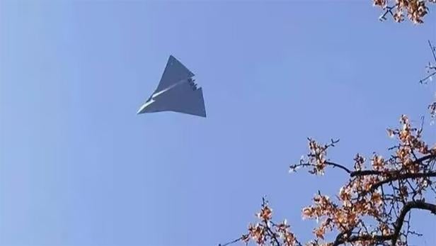 Dreieckiges Tarnkappenflugzeug fliegt am klaren Himmel, Äste mit Blüten im Vordergrund.