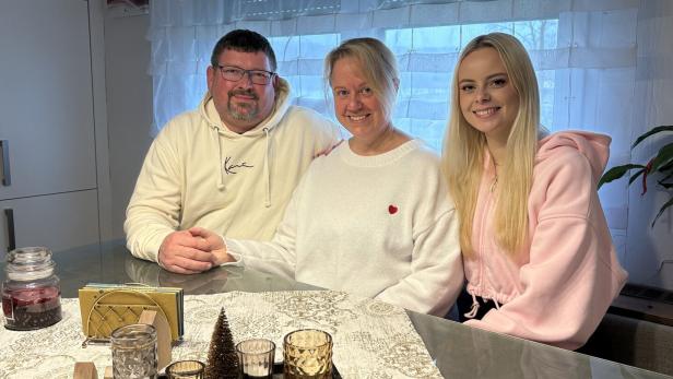 Drei Personen sitzen lächelnd an einem Tisch mit Kerzen und Dekoration, im Hintergrund ein Fenster mit Gardinen.
