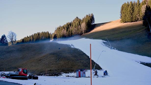 Eine schmale Skipiste mit Kunstschnee verläuft durch eine grüne Wiese, umgeben von Nadelbäumen und Schneekanonen.