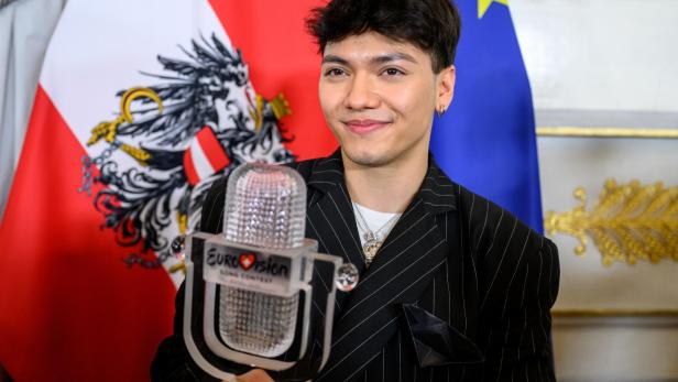 Ein junger Mann hält eine Eurovision-Trophäe vor österreichischer und EU-Flagge und lächelt.