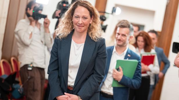 Eine Frau im Anzug geht lächelnd durch einen Flur, gefolgt von mehreren Personen mit Aktenmappen und umgeben von Fotografen.