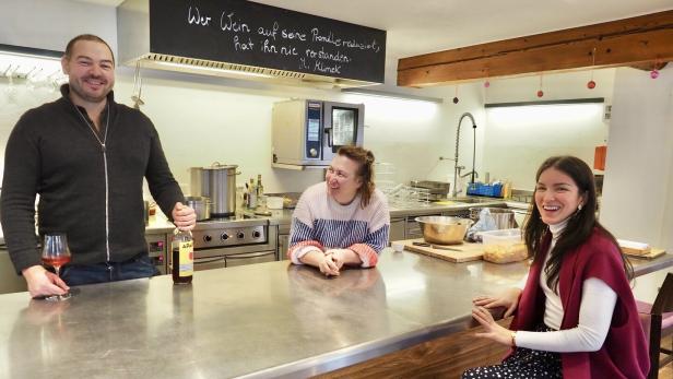 Drei Personen stehen und sitzen lächelnd an einer Küchentheke mit Getränken.