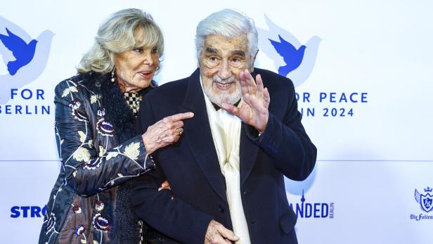 Ein älteres Paar steht lächelnd nebeneinander, während der Mann winkt und die Frau ihn am Arm hält.