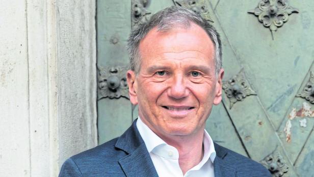Ein Mann mit grauen Haaren trägt ein dunkelblaues Sakko und ein weißes Hemd und steht vor einer verzierten Tür.