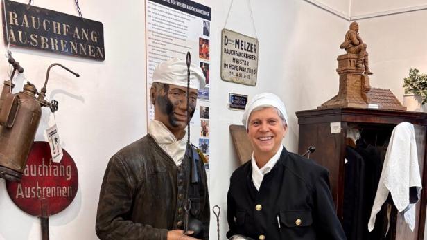 Eine Person in traditioneller Schornsteinfeger-Uniform steht lächelnd neben einer Schornsteinfeger-Figur mit Werkzeugen.