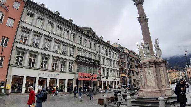 Eine belebte Fußgängerzone mit historischen Gebäuden, einer hohen Säule mit Statuen und mehreren Passanten bei bewölktem Himmel.