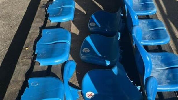 Mehrere blaue Kunststoffsitze in einer Stadionreihe sind beschädigt oder fehlen teilweise.