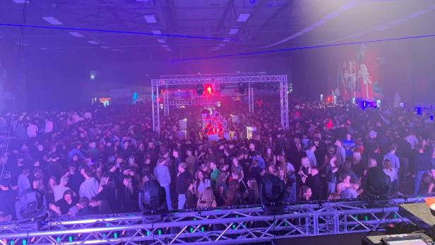 Große Menschenmenge feiert in einer Halle mit bunter Beleuchtung und Musikbühne.