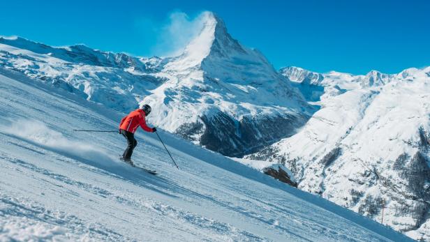 Eine Person fährt auf Skiern eine verschneite Piste hinab, im Hintergrund ragt das Matterhorn in den blauen Himmel.