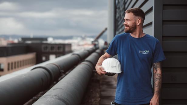 Mitarbeiter mit immOH!-Shirt hält Schutzhelm auf einem Gebäudedach neben großen Rohrleitungen.
