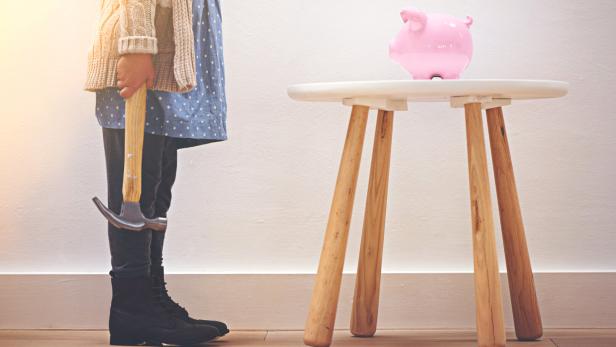 Ein Kind hält einen Hammer in der Hand und steht vor einem Tisch, auf dem ein rosa Sparschwein steht.