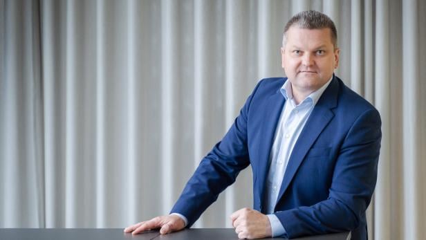 Ein Mann im blauen Anzug steht an einem Tisch vor einem hellen Vorhang und blickt ernst in die Kamera.